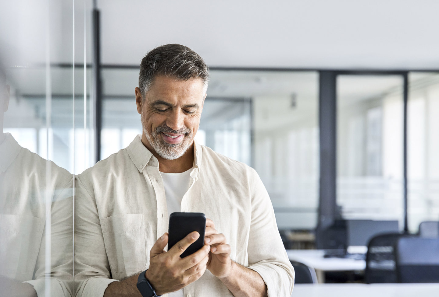 En mand står på et kontor og smiler, mens han kigger på sin smartphone; han har en lys skjorte og et smartur på, og i baggrunden ses glasvægge.