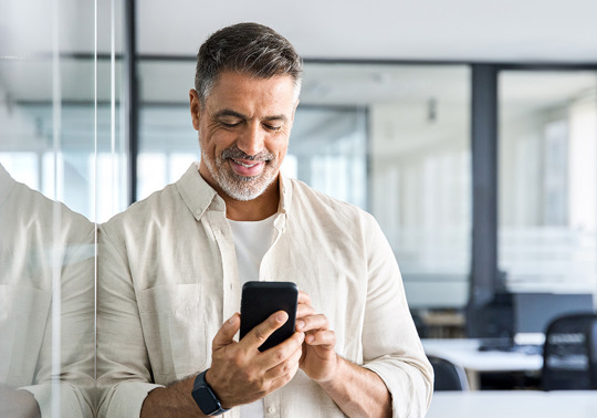 Smilende mand, der bruger en smartphone på et moderne kontor, stående og lænet op ad en glasvæg.