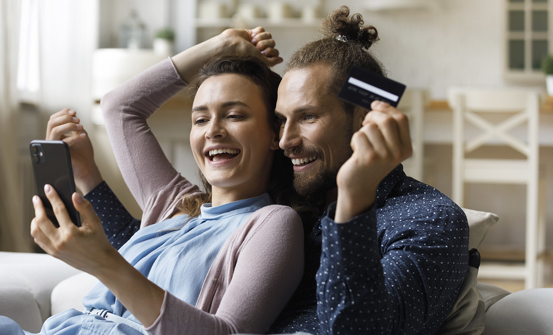 Et smilende par sidder i en sofa med en smartphone og et kreditkort i hånden og ser ud til at foretage et onlinekøb sammen.