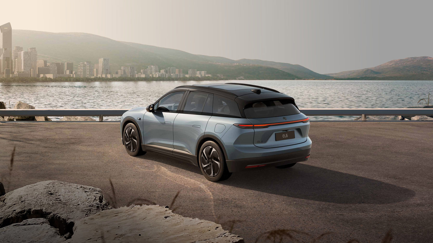 En blå elektrisk SUV holder parkeret ved havnefronten, med byens skyline og bakker i baggrunden under en klar himmel.